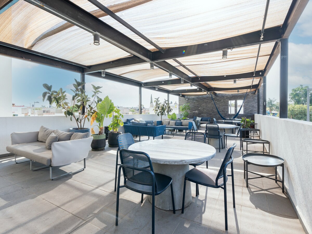 A spacious terrace is featured, furnished with a round table surrounded by various seating options, including lounge chairs and modern outdoor sofas. Sunlight filters through a retractable canopy, while potted plants add greenery to the setting, creating an inviting atmosphere suitable for relaxation.