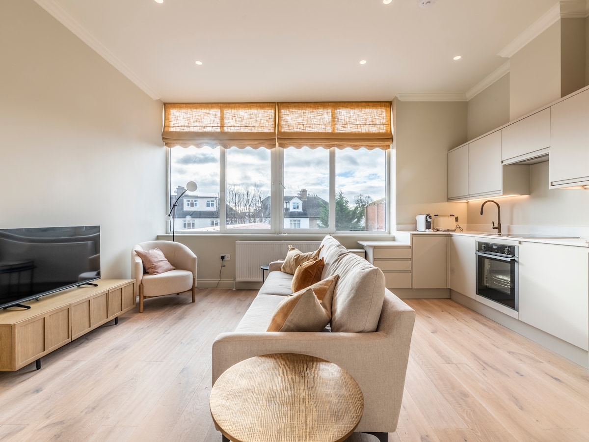 A spacious living area features a comfortable beige sofa, a wooden coffee table, and a light armchair. Large windows allow natural light to fill the space, with soft fabric shades providing aesthetic appeal. The modern kitchen is visible, showcasing high-end appliances and ample counter space.