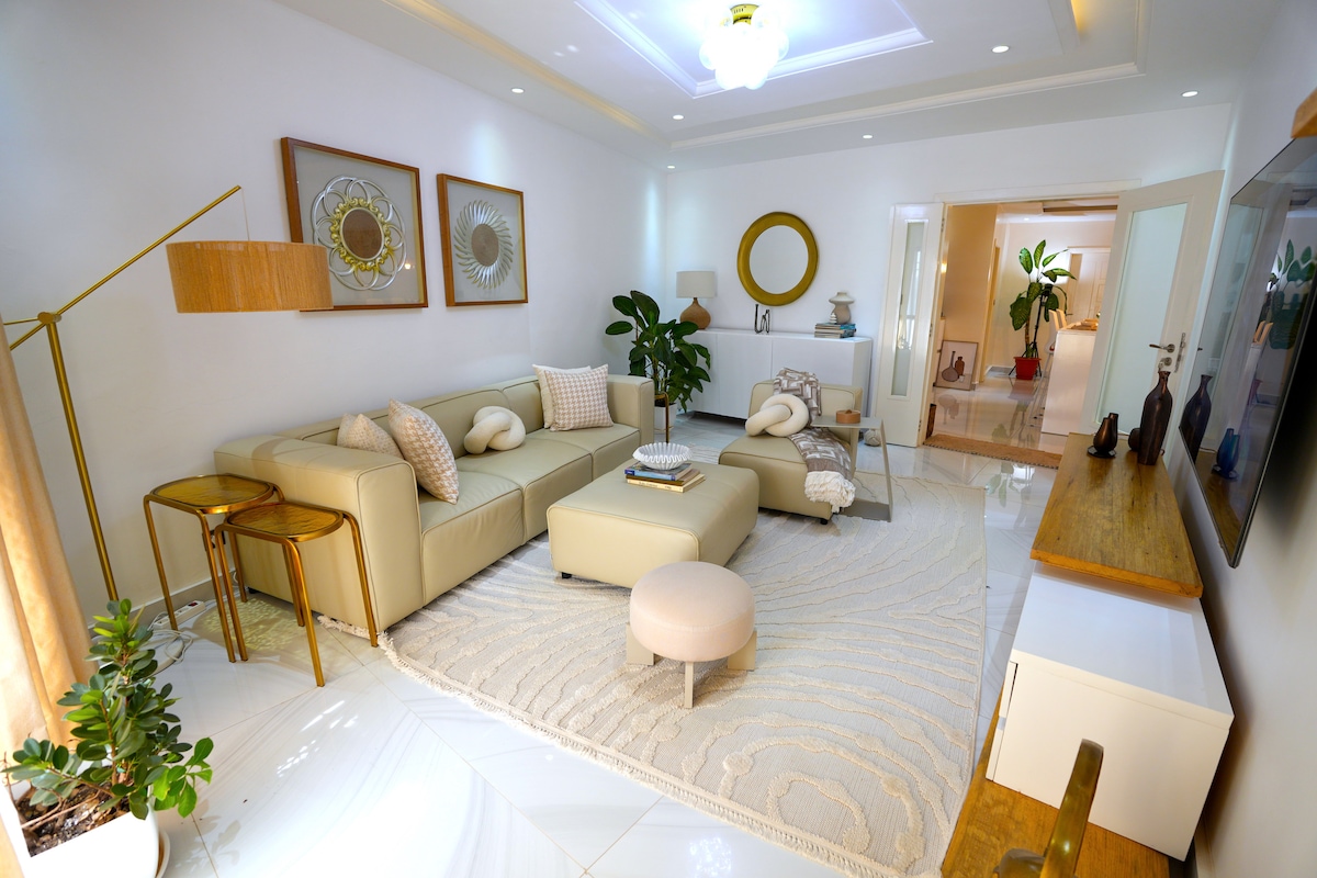A modern living area is presented with a light-colored sectional sofa and matching ottoman. A plush rug lays beneath a glass coffee table, and decorative plants enhance the space. Wall art and mirrors add visual interest, while soft lighting creates a warm ambiance.
