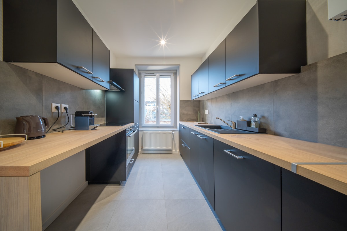 The kitchen features sleek, modern cabinetry in dark tones with integrated handles. Natural light floods through a window above the sink, illuminating the spacious countertops. Appliances, including a kettle and toaster, are visible, alongside a minimalist design that emphasizes functionality.