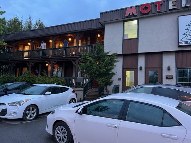 A two-story building with a wooden balcony features multiple entrances, visible on the upper level. The ground is occupied by several parked cars, while lush greenery is present along the sides. The overall aesthetic reflects a classic motel style.