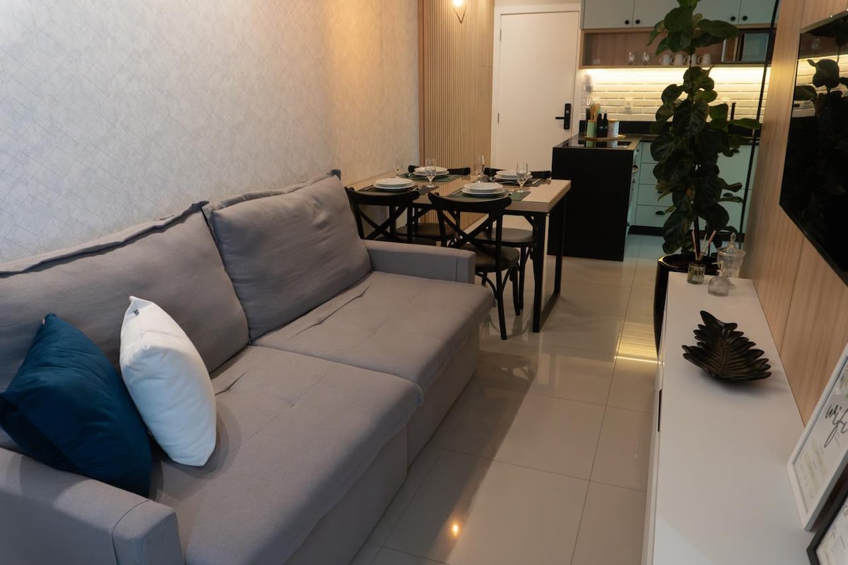 A comfortable living area is featured with a light gray sofa, complemented by decorative cushions. A dining table for four is prepared nearby, with stylish table settings in place. The kitchen, visible in the background, offers modern cabinetry and appliances.