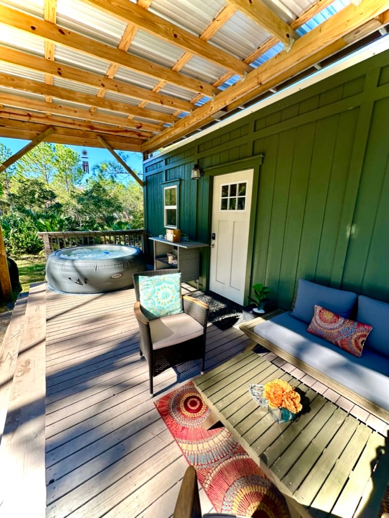 An outdoor living space features a covered deck with a hot tub and seating area. A small table is accompanied by two chairs, while a plush sofa rests against the wall. Decorative cushions and a vibrant rug add color to the neutral tones of the furniture.
