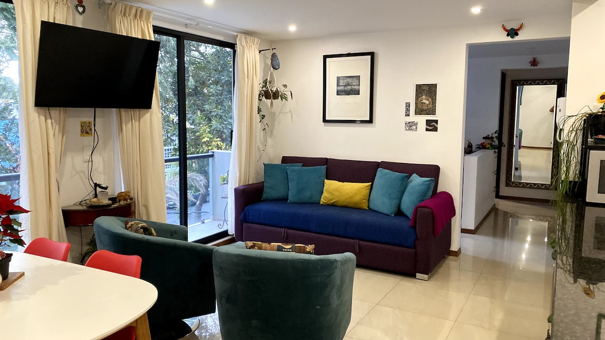 A well-lit living area features a purple sectional sofa adorned with yellow and pink pillows. Nearby, a dining table with red chairs is positioned next to a large window, which offers views of the surrounding greenery. Wall art and indoor plants enhance the inviting atmosphere.