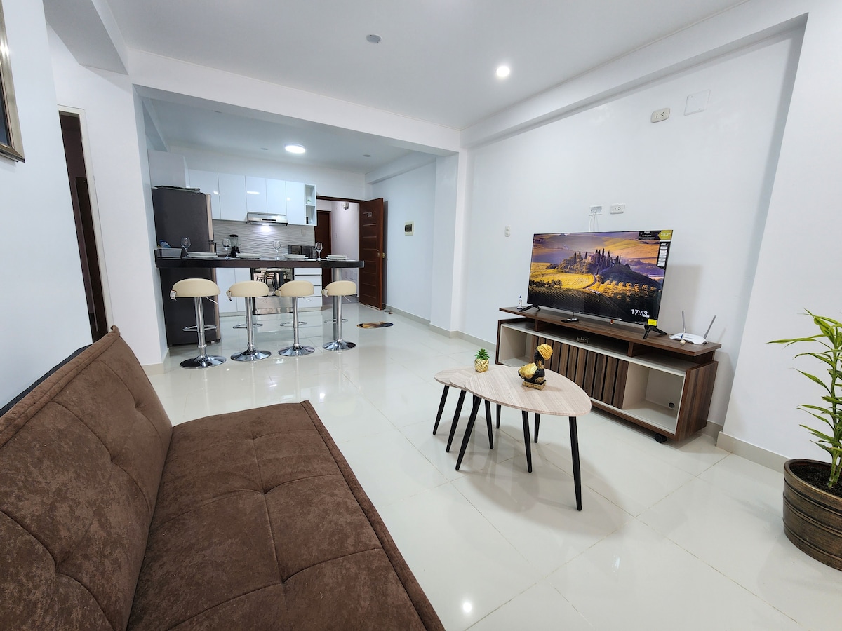 A bright living area showcases a comfortable brown sofa opposite a modern entertainment center. A flat-screen television rests on a white and wood media console. A small round coffee table is positioned in front of the sofa, surrounded by a potted plant and a spacious kitchen area visible in the background.
