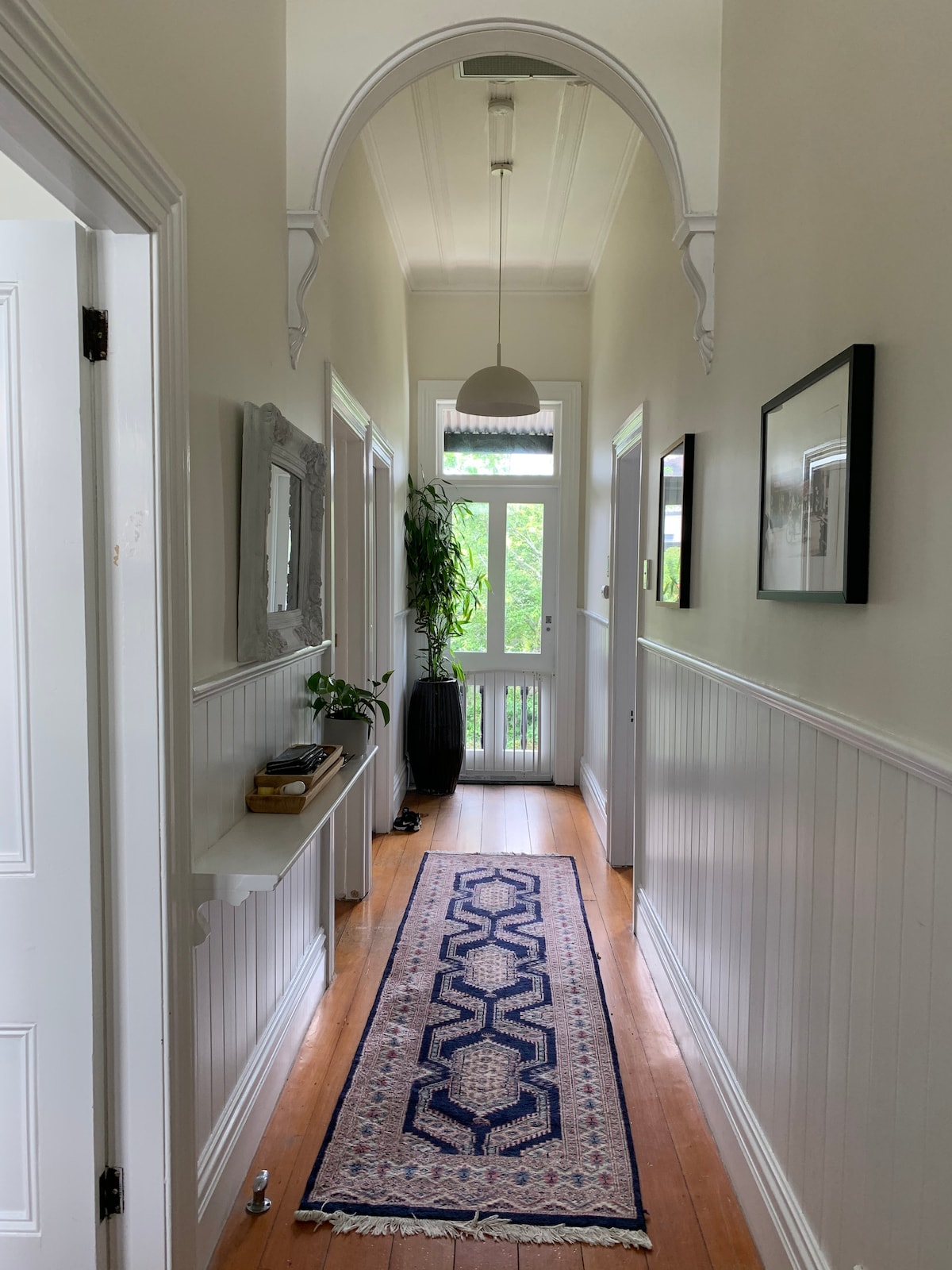 A hallway features wooden flooring and a patterned runner. Decorative elements include framed artwork on the walls and a small console table with plants. Natural light enters through the windows at the end of the corridor, highlighting the high ceilings and archway.