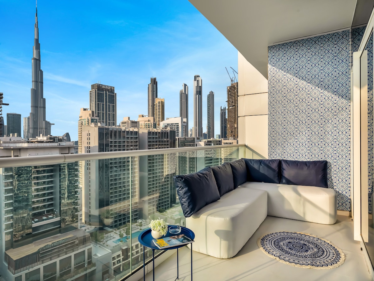 A cozy balcony with a curved white sofa provides comfortable seating, adorned with navy blue cushions. A small table with a decorative plant sits nearby, while the panoramic view showcases the iconic Burj Khalifa and Dubai's impressive skyline in the background under a clear blue sky.