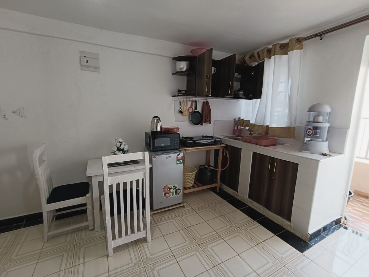 A compact kitchen area is presented with a small dining table and two chairs. The kitchen features cabinetry, a stove, and a refrigerator. Natural light enters through a window covered with sheer curtains, illuminating the tiled floor.