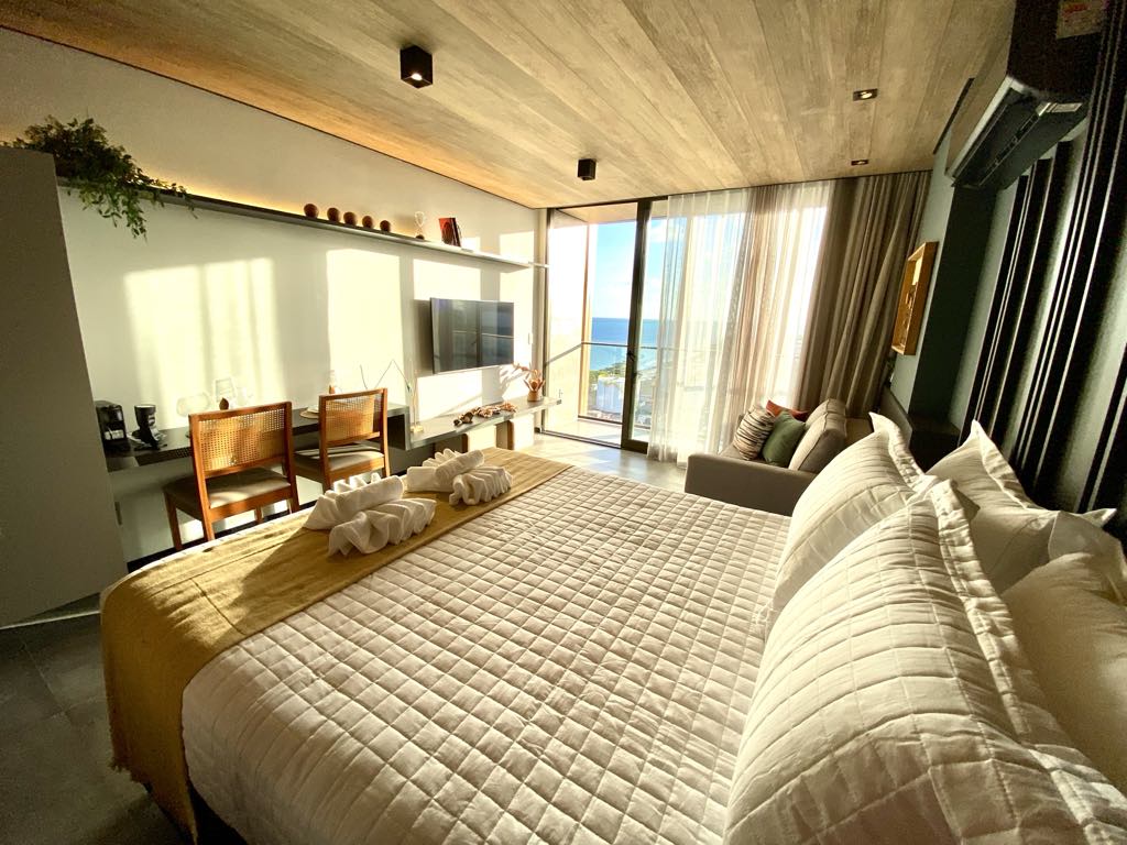 A modern studio space features a spacious bed with neatly arranged white linens. A cozy seating area and dining space are visible. Natural light filters through large glass doors, revealing a view of the sea. The wooden ceiling and sleek furnishings add to the contemporary design.