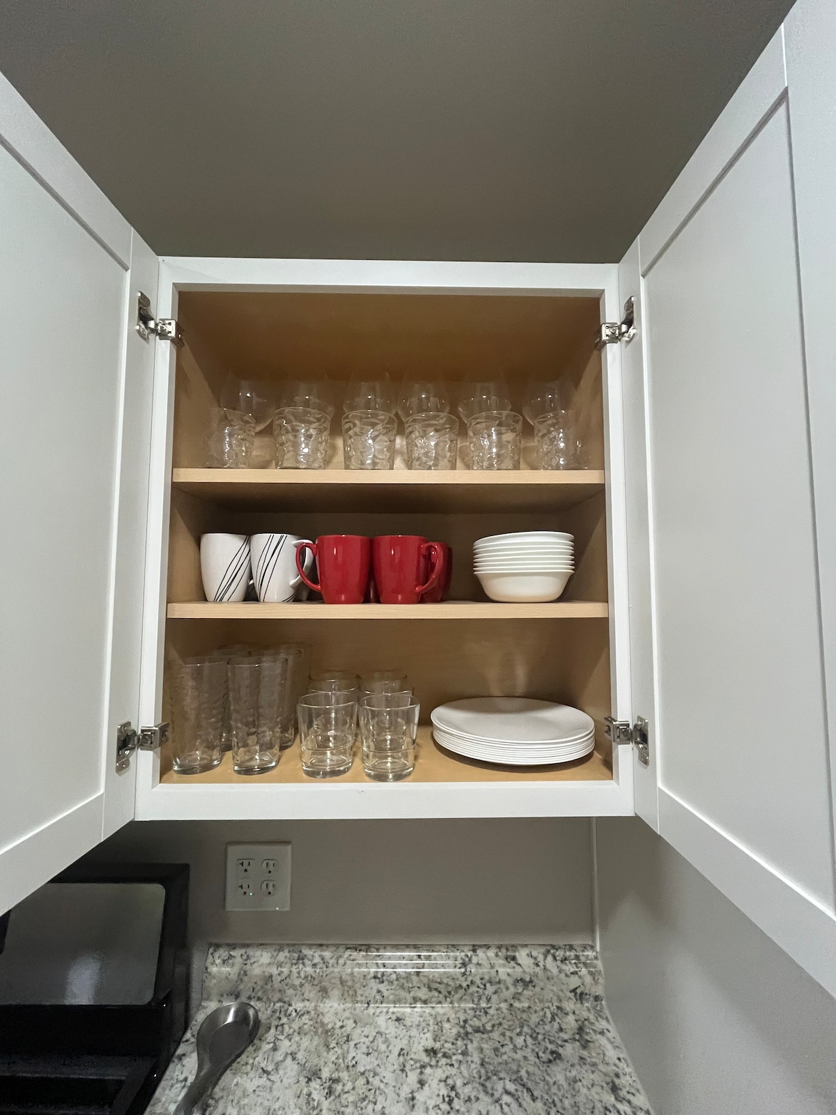 A kitchen cabinet is open to reveal organized dishware. The top shelf contains clear glassware and mugs, while the lower shelf features white plates and red mugs. The cabinet's interior is light-colored, contrasting with the dark granite countertop below.