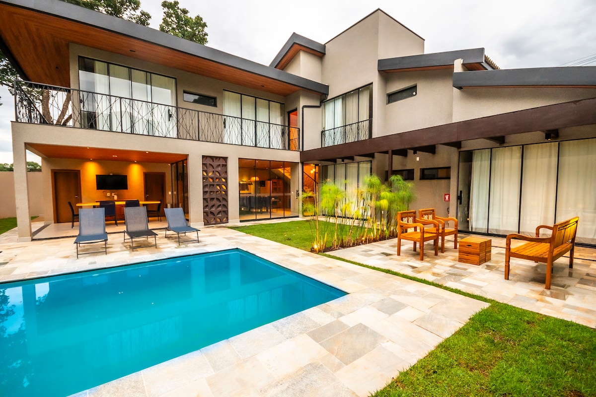 The modern exterior showcases a private pool surrounded by lounge chairs and a well-maintained garden. Large glass windows allow natural light to fill the living area, while wood accents add a touch of warmth to the contemporary design.