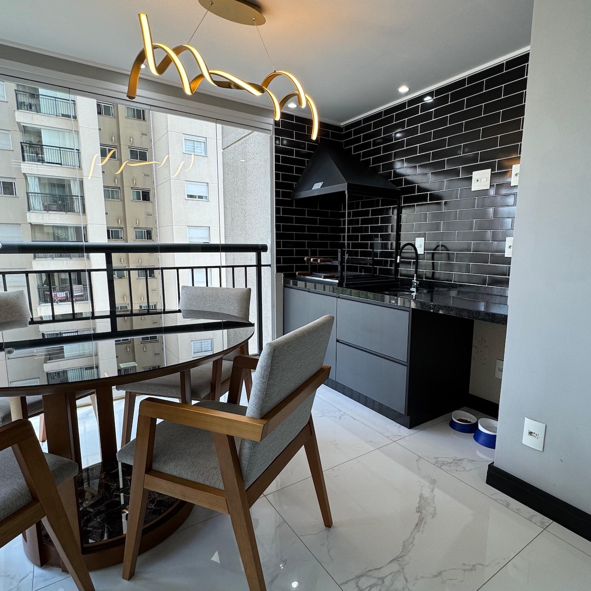 A modern kitchen area is seen with sleek black backsplash tiles that contrast with light cabinetry. A dining table with four light grey chairs is positioned near a glass railing, providing a view of outside. Natural light enhances the airy atmosphere.