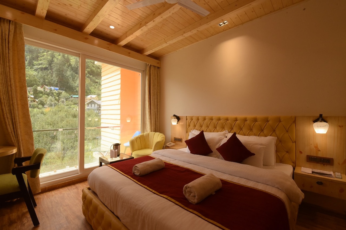 A well-appointed bedroom features a spacious bed with decorative pillows and soft linens. Large glass doors provide natural light and offer a view of the surroundings. A cozy seating area with a bright chair complements the warm wood ceiling and flooring.