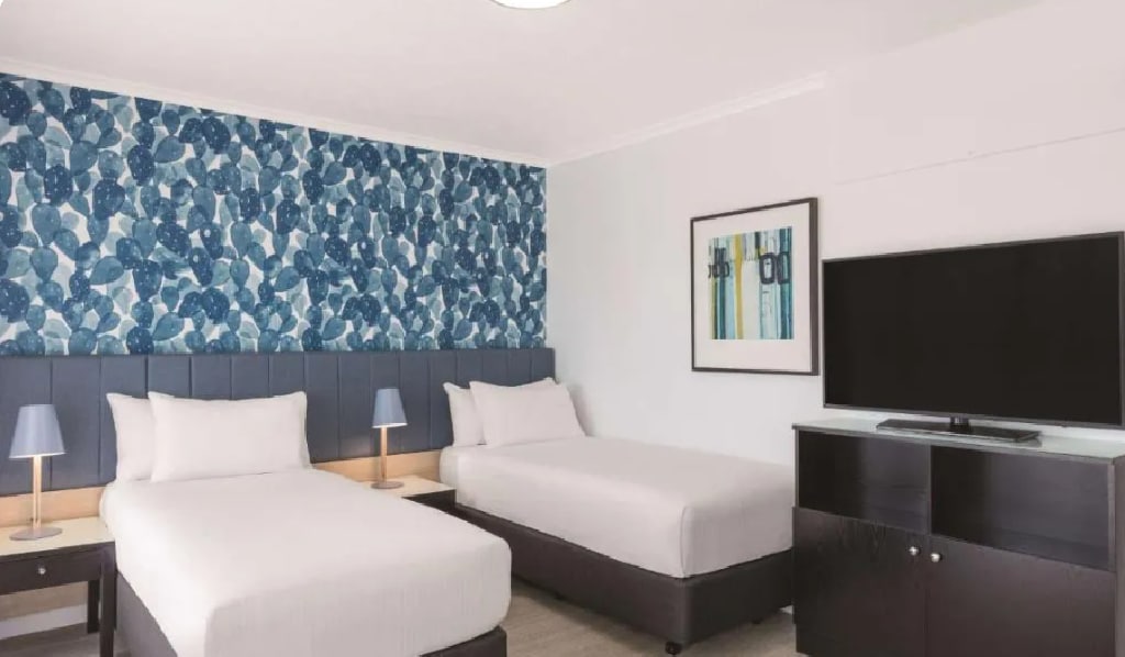 A well-lit guest room features two spacious beds with white linens. A decorative blue and white patterned wall enhances the modern design. Side tables with lamps are positioned beside each bed, while a flat-screen TV is mounted on a cabinet opposite.