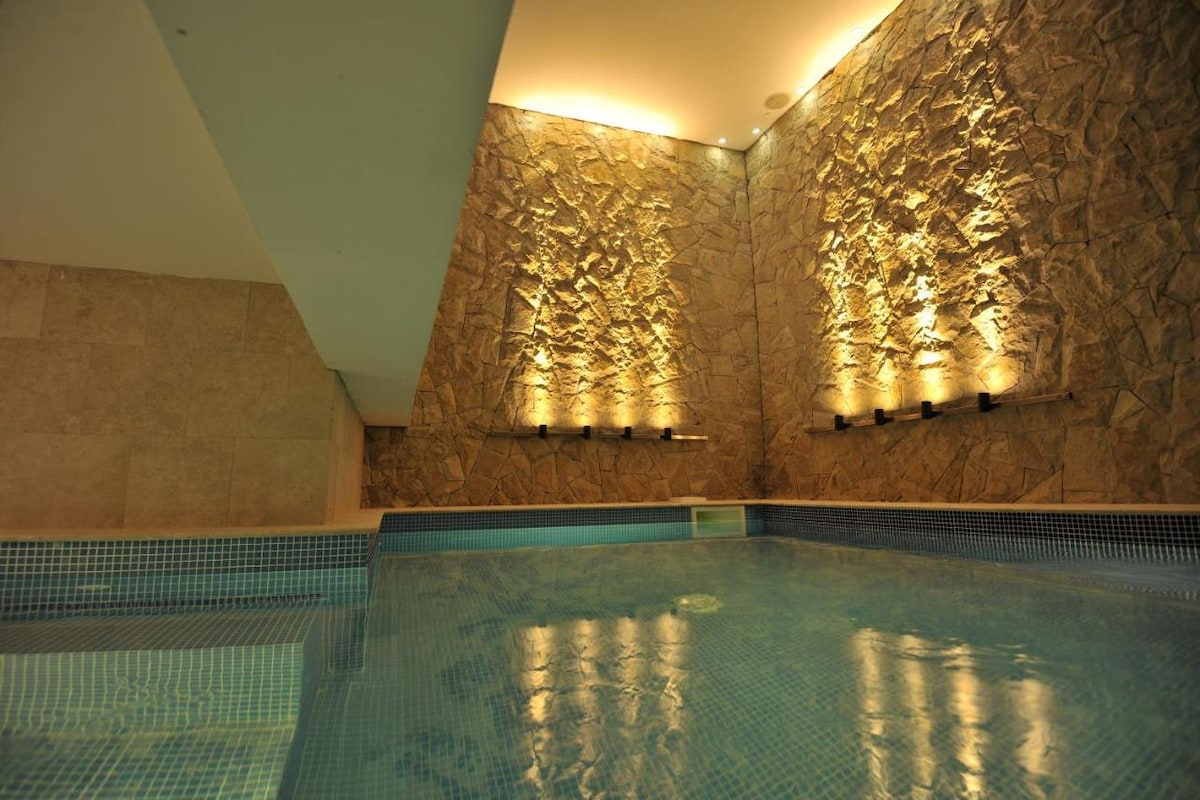 An indoor pool features smooth, blue tiles with shimmering water under gentle lighting. The stone wall is highlighted with soft lighting, creating a serene atmosphere. The design emphasizes clean lines and a tranquil setting, perfect for relaxation.
