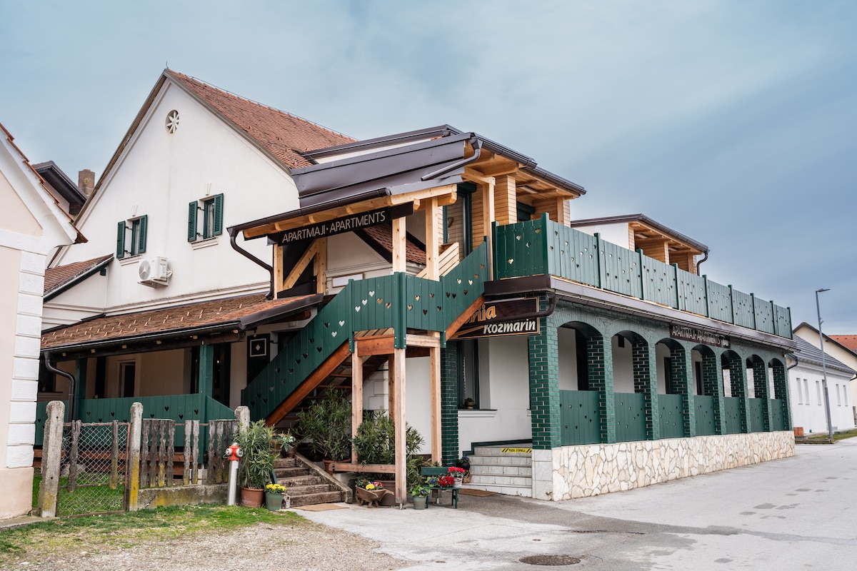 The exterior of Apartments Babi features a modern, multi-level structure with a wooden staircase leading to a covered balcony. Green railings and flower planters add a touch of charm to the inviting entrance. The building is set against a soft, overcast sky.