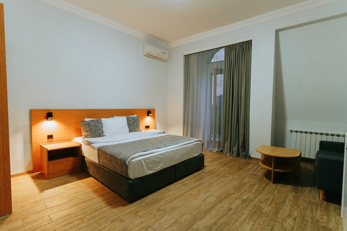 A bright and spacious bedroom features a large bed with soft bedding, flanked by two bedside lamps. Natural light filters through sheer curtains covering a large window. A wooden table and two chairs are positioned in one corner, complemented by a cozy seating area.