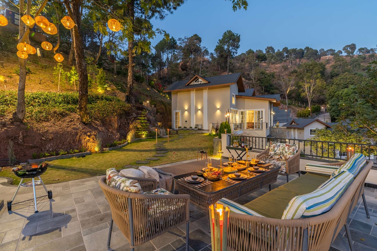 An inviting outdoor seating area is featured, surrounded by lush greenery and gently lit by hanging lanterns. A dining setup is arranged on a stone patio, complemented by a BBQ grill nearby. The villa's modern architecture stands elegantly in the background.
