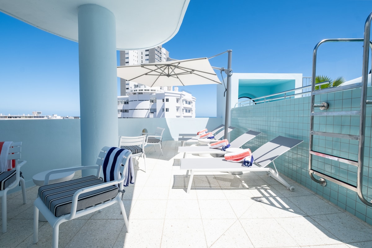 A spacious rooftop pool area features several lounge chairs arranged neatly under a large umbrella. Bright blue walls and tiles create a fresh ambiance, while clear sky and city buildings provide a vibrant backdrop. The area is designed for relaxation and enjoyment.
