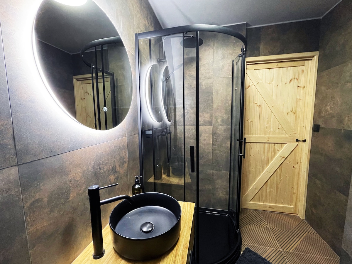 A modern bathroom features a round mirror illuminated by soft lighting. A sleek, dark circular sink sits atop a wooden vanity. A curved glass shower enclosure is visible, and a wooden door adds a natural touch to the space.