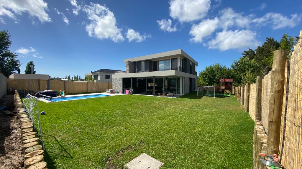 A modern house with large glass windows overlooks a spacious green yard. A fenced area contains a heated pool, and a small playhouse is visible nearby. The yard offers ample space for outdoor activities, enhanced by the surrounding trees and blue sky.