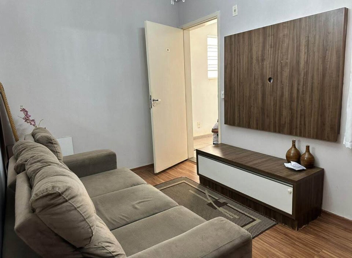 A living area features a plush, L-shaped sofa positioned against a light-colored wall. A wooden media console with decorative vases is situated beneath a wall-mounted flat-screen. A beige area rug is placed on a laminate floor, complementing the neutral color scheme.