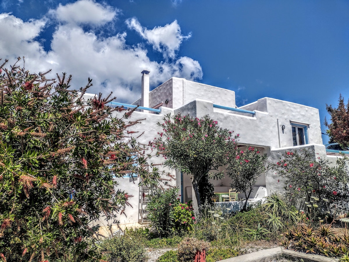A traditional whitewashed house is surrounded by vibrant greenery and flowering plants. The structure features a chimney and two balconies, contributing to its simple Cycladic style. Blue skies and soft clouds create a bright atmosphere above the home.