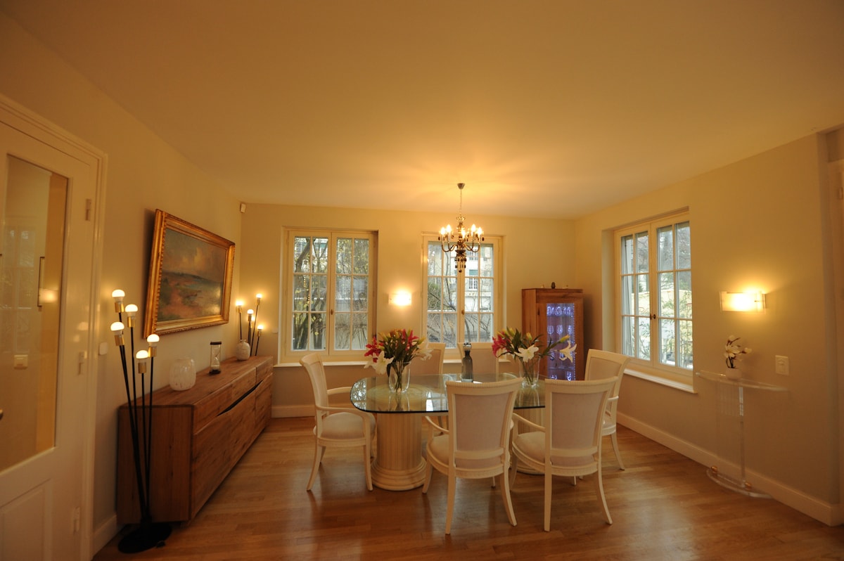 A dining area is revealed with a round glass table surrounded by six white chairs. Soft light is provided by a chandelier and several standing lamps. A wooden sideboard is positioned against the wall, and large windows allow natural light to fill the space.
