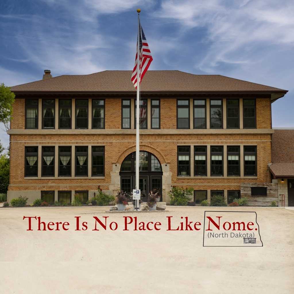 The exterior of a historic school building is showcased, featuring large windows framed by brickwork. An American flag is displayed prominently in front, and the entrance is welcoming with a landscaped path leading towards it. The setting reflects the building's educational heritage.