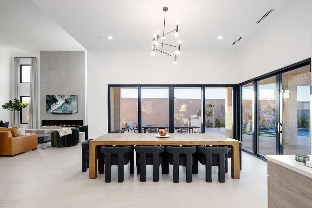 A spacious dining area features a large wooden table surrounded by dark, textured chairs. Floor-to-ceiling windows overlook the backyard, allowing natural light to fill the room. A modern chandelier hangs above the table, while a cozy seating area is visible in the background.