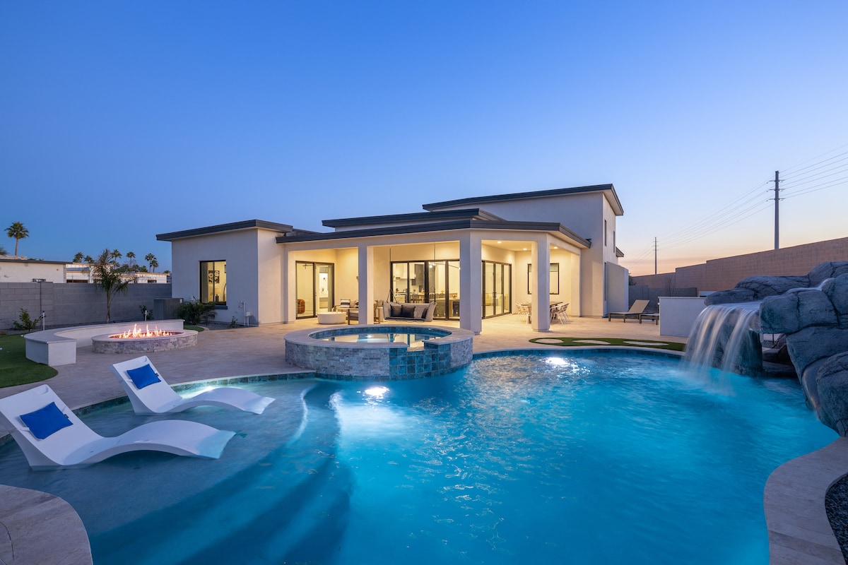 A modern home with expansive glass doors opens to a spacious outdoor area. A large pool features a soothing waterfall and an attached hot tub. Comfortable loungers sit beside the pool, and the scene is softly illuminated by the twilight sky.