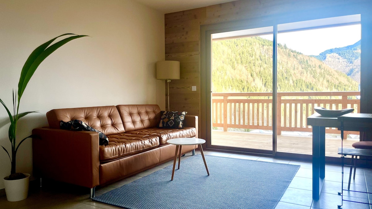 A spacious living area presents a comfortable brown leather sofa, accompanied by a small coffee table. Large sliding glass doors offer a picturesque view of the surrounding mountains and provide access to the terrace. A decorative plant adds a natural touch to the room.