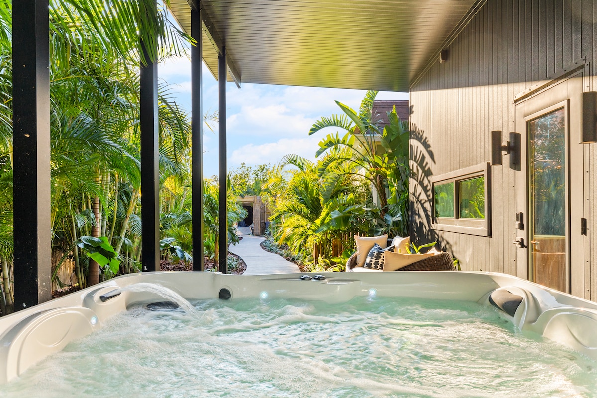 A private hot tub, filled with bubbling water, is surrounded by lush tropical landscaping. The space features a covered patio area that provides shade. A winding pathway can be seen in the background, leading through the vibrant greenery.