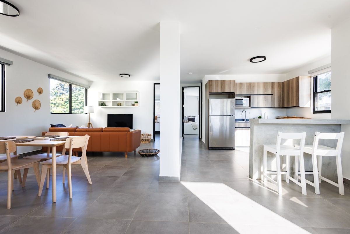 An open-plan living area is showcased, featuring a light-colored sofa paired with wooden dining furniture. A modern kitchen is visible, equipped with stainless steel appliances and ample counter space. Natural light fills the room through multiple windows, highlighting the clean lines and contemporary design.
