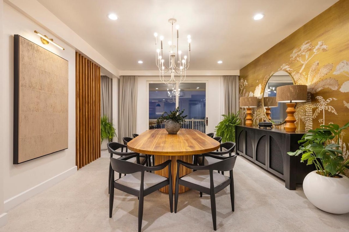 A dining area features a large round wooden table surrounded by sleek chairs. A statement chandelier hangs above, providing soft lighting. A sideboard displays decorative pieces, while accent plants add a touch of greenery. Large windows offer views of the surroundings through elegant drapes.