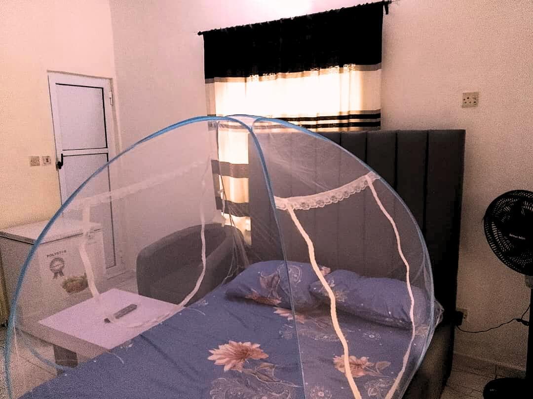 A sleeping area is defined by a spacious bed covered with light bedding featuring floral patterns. A protective netting is draped over the bed. Nearby, a small table holds a remote control, with a fan positioned beside a cozy gray sofa. Natural light filters through the partially covered window.