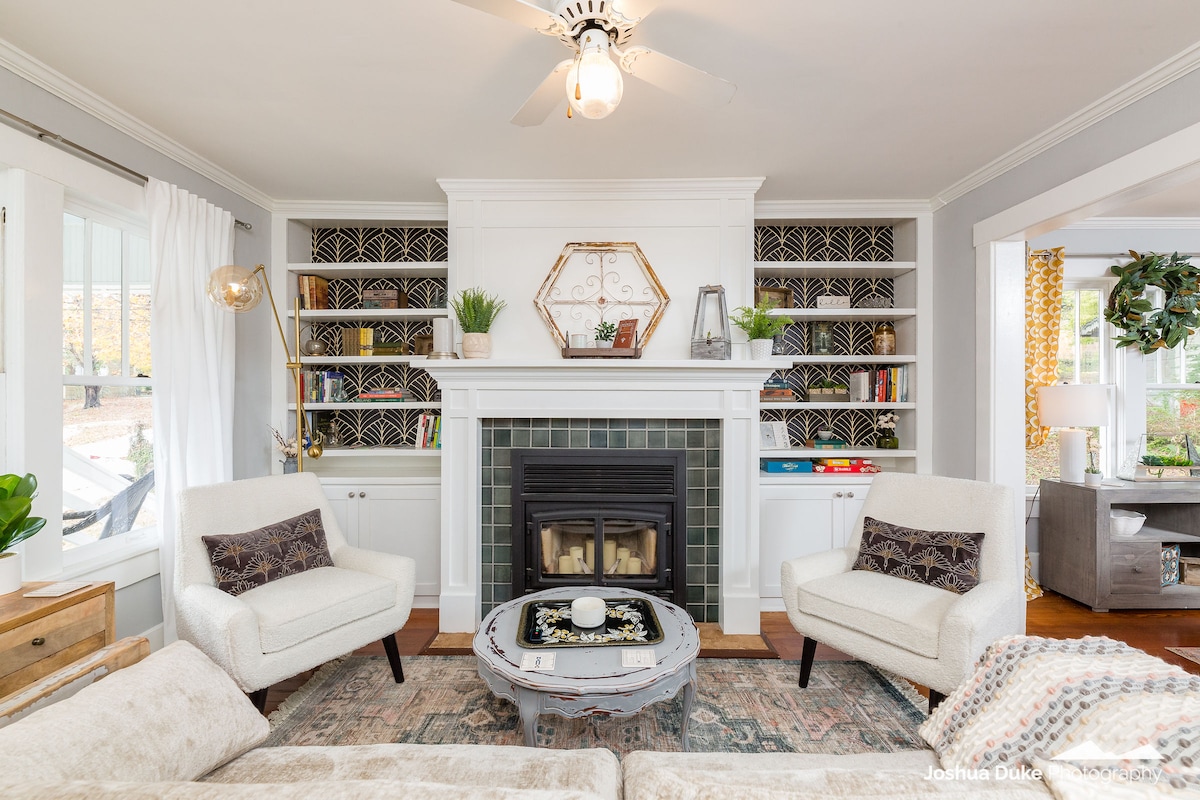 A cozy living area is highlighted by a central fireplace set within a white mantel. Flanking the fireplace are built-in shelves displaying plants and books. Two armchairs provide comfortable seating, while a patterned rug and a decorative table anchor the space.