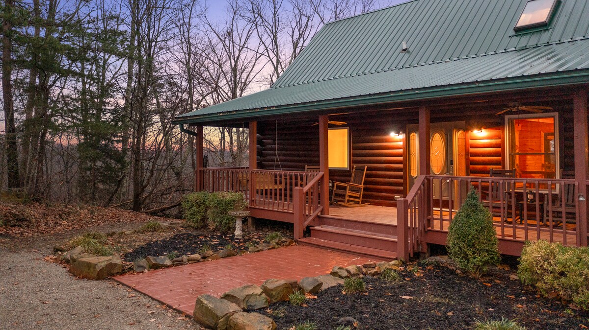 The cabin's exterior showcases a rustic wooden structure with a green metal roof. A covered porch features rocking chairs and is surrounded by landscaped gardens. Pathway stones lead to the entrance, set against a backdrop of trees as dusk settles.
