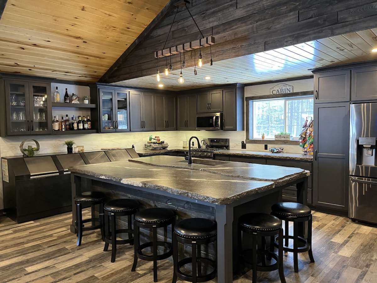 A modern kitchen is highlighted by a large central island with a dark countertop and seating for several guests. Dark cabinets provide ample storage, while stainless steel appliances and under-cabinet lighting enhance functionality. The wooden ceiling adds warmth to the space.