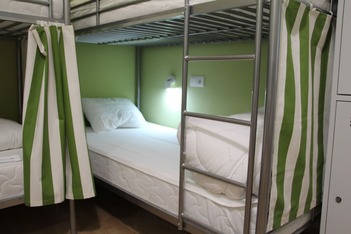 A shared dormitory room features metal bunk beds with neatly made beds, each equipped with white linens. Green striped curtains provide privacy at the side of the lower bunk. A soft light is positioned above the upper bed, illuminating the space.