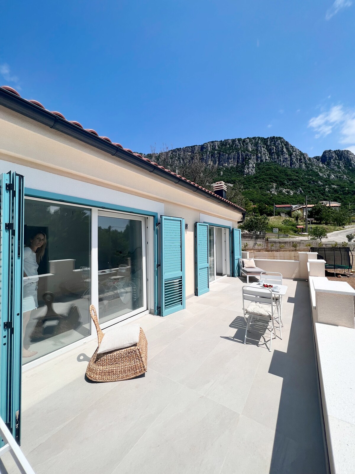 A bright outdoor terrace features light-colored tiled flooring and is equipped with a small dining table and chairs. Blue shutters frame the large glass doors that open to the interior. Lush green mountains provide a scenic backdrop under a clear blue sky.