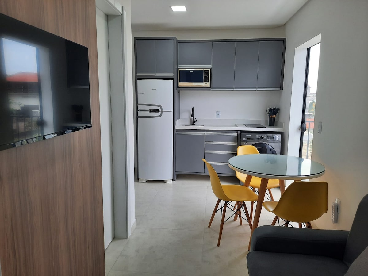 A modern kitchen area is shown, featuring sleek gray cabinetry and stainless steel appliances. A round glass dining table is accompanied by four yellow chairs. Light enters through a large window, illuminating the spacious layout, while a wall-mounted TV adds convenience.