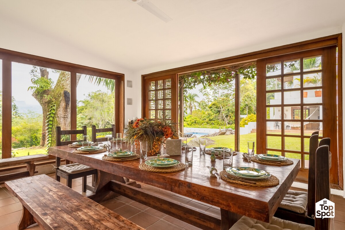 A spacious dining area is seen with a large wooden table set for a meal. Natural light pours in through expansive glass doors, revealing views of lush gardens and the private pool outside. Simple wooden benches are provided for seating.