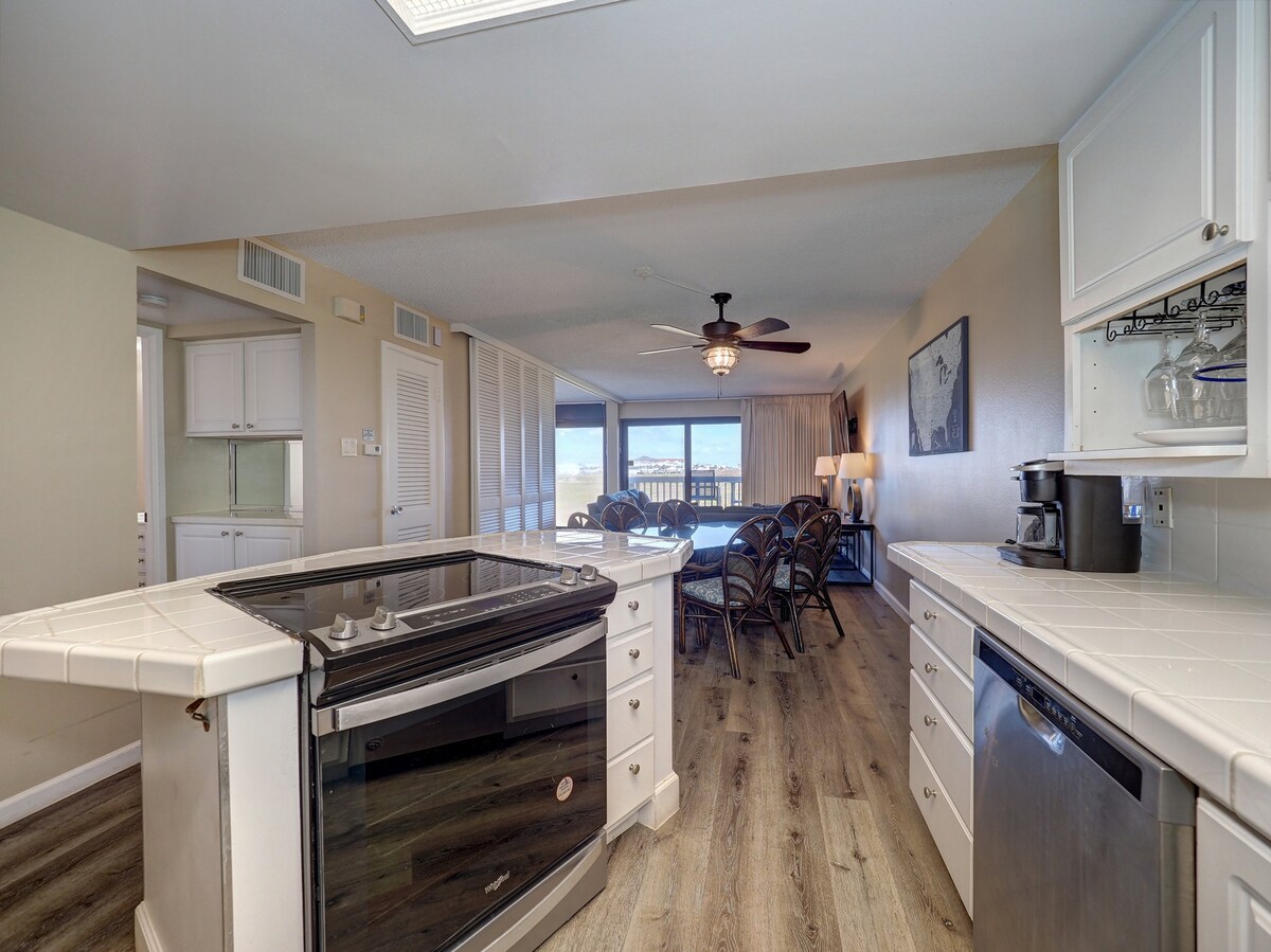 An open kitchen area features a modern island with a stovetop and sleek cabinets. A dining space with a round table and woven chairs offers ocean views through large sliding glass doors. Natural light fills the room, enhancing the overall bright atmosphere.