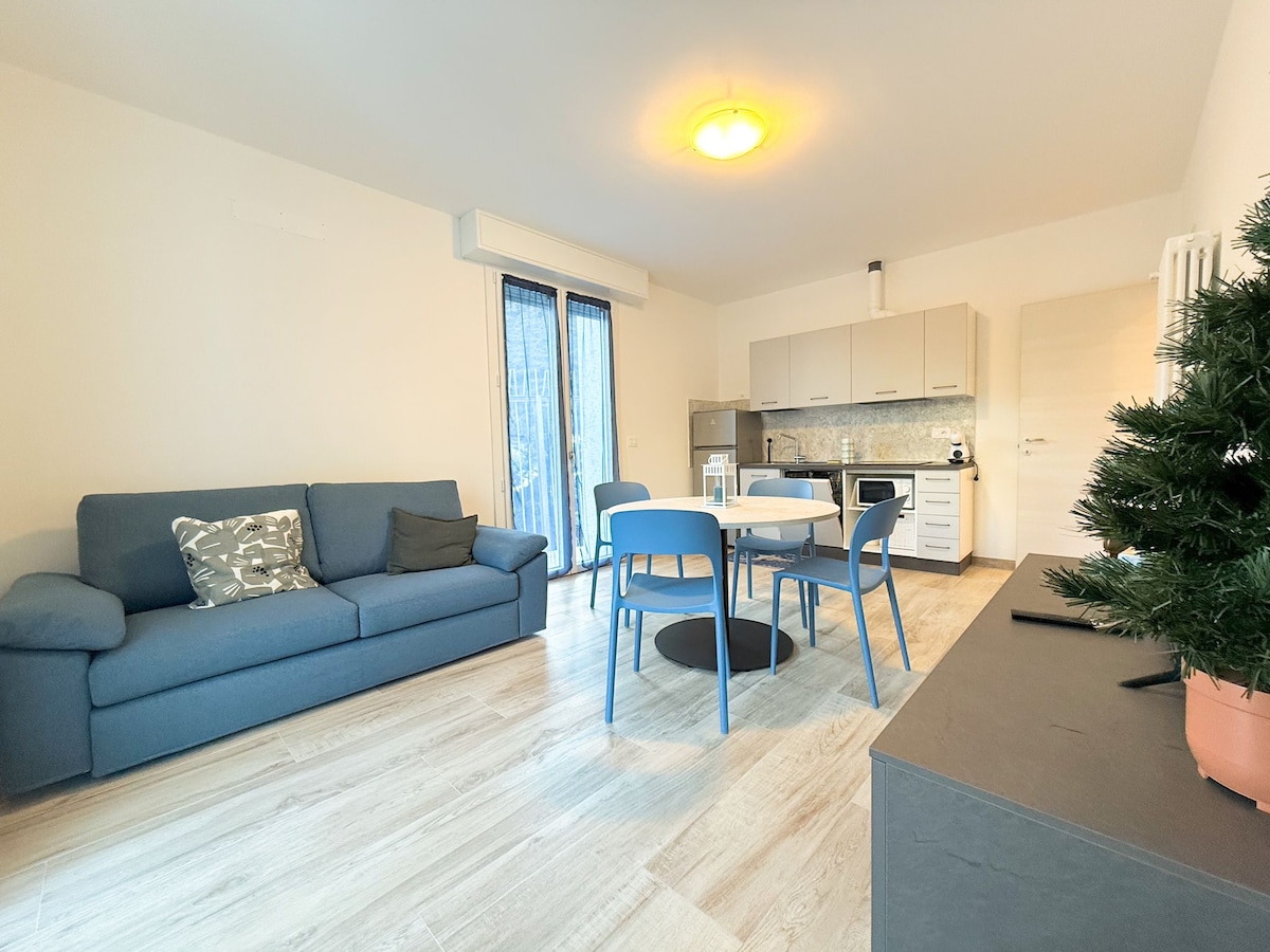 A furnished living space is displayed, featuring a blue sofa and a round dining table surrounded by four blue chairs. The kitchen area is visible in the background, equipped with modern appliances. Large windows allow natural light to brighten the room.