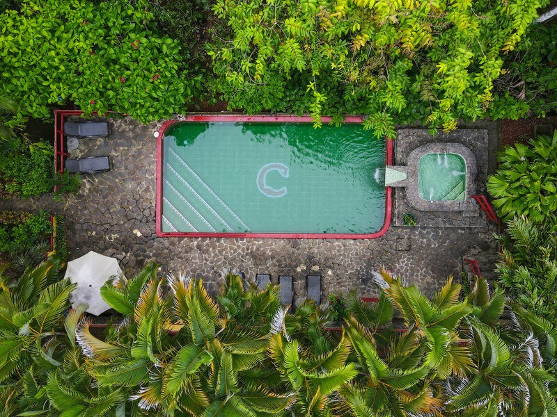 An aerial view captures a refreshing swimming pool surrounded by lush tropical greenery. Stone pathways and lounge chairs are positioned around the pool area, while a small hot tub is visible alongside it, inviting relaxation amid the vibrant flora.