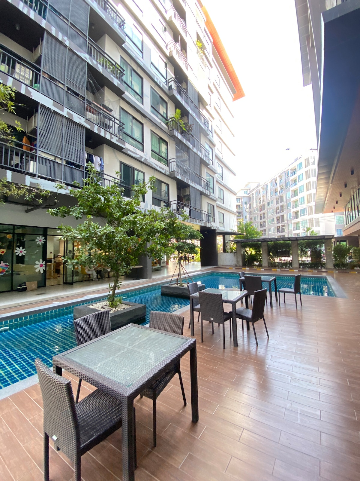 An outdoor area is presented, featuring multiple tables surrounded by chairs under the shade of trees. A swimming pool with clear blue water is visible, with modern buildings bordering the space. Well-maintained tiles enhance the overall clean and organized appearance.