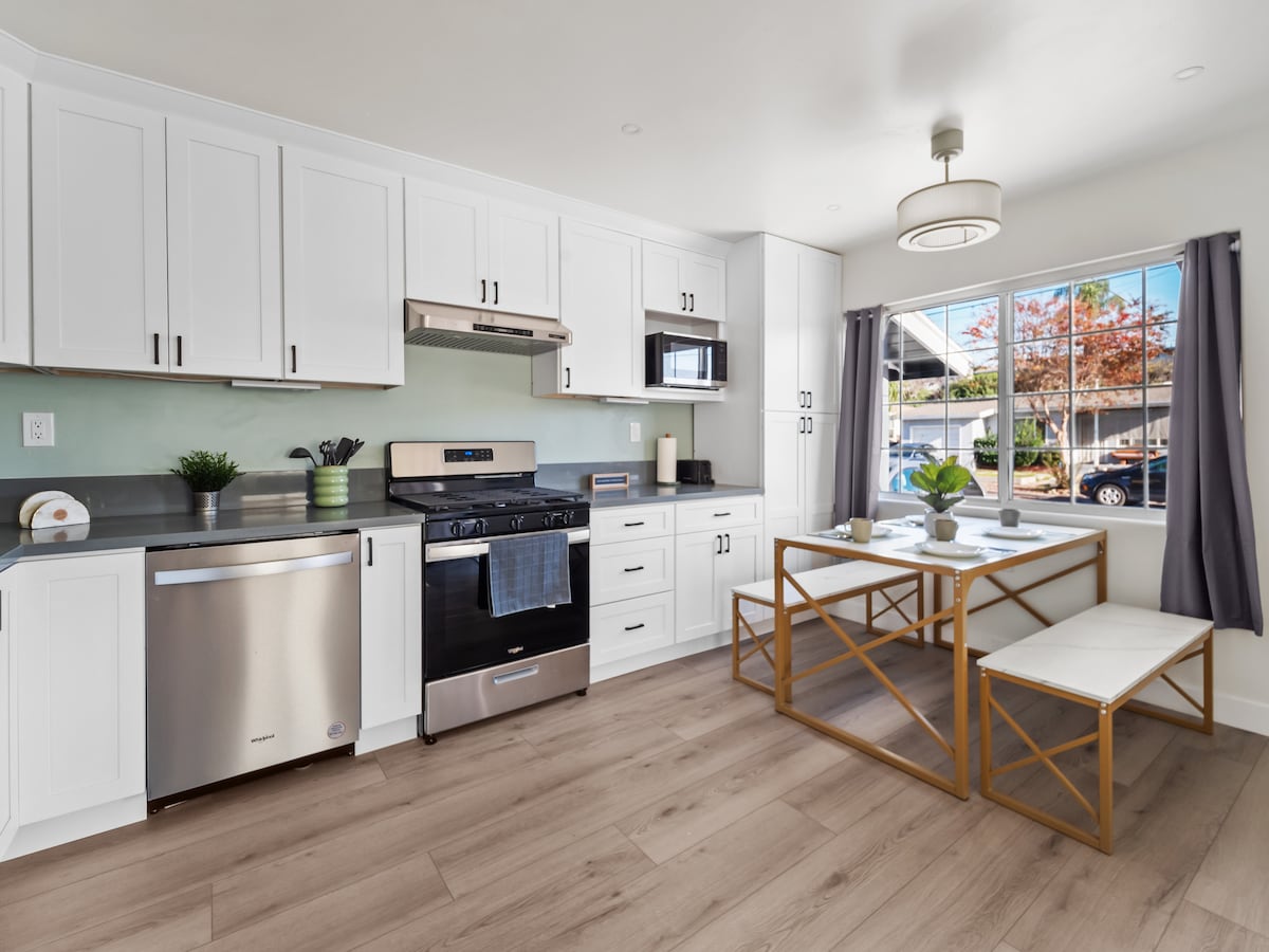 A modern kitchen features white cabinetry and stainless steel appliances, including a stove and dishwasher. A dining area with a rectangular table and two benches is illuminated by natural light from a nearby window, while light wood flooring adds warmth to the space.
