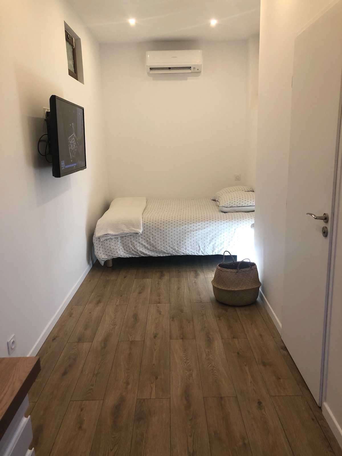 A compact studio features a spacious bed covered with a dotted white duvet, positioned against a light-colored wall. A television is mounted on the wall opposite the bed. A small basket sits on the floor, and an air conditioning unit is visible above the bed.