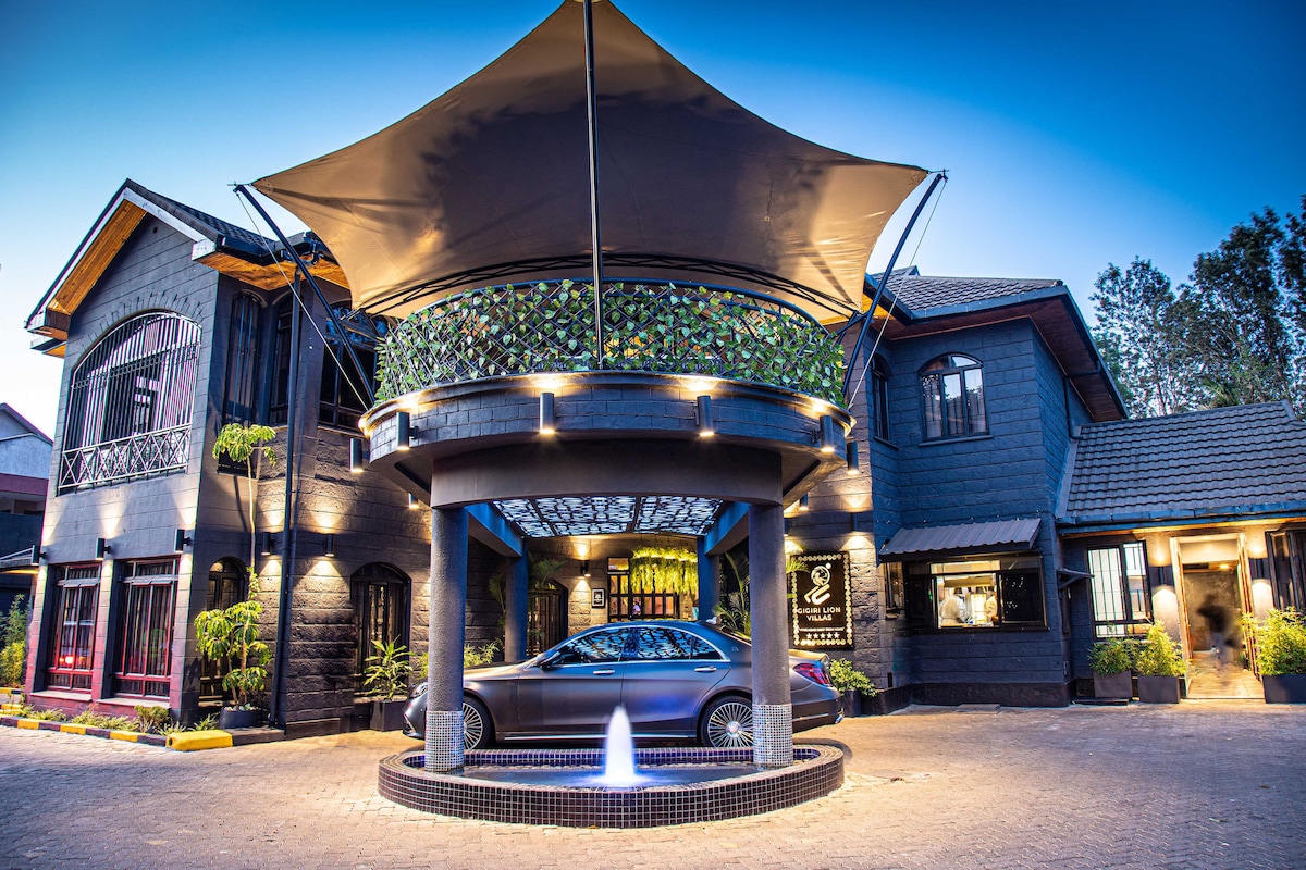 The entrance features a striking architectural canopy supported by a circular structure. A sleek car is positioned near a small fountain, with landscaped greenery visible around the entrance. The building showcases a harmonious blend of modern and traditional design elements.
